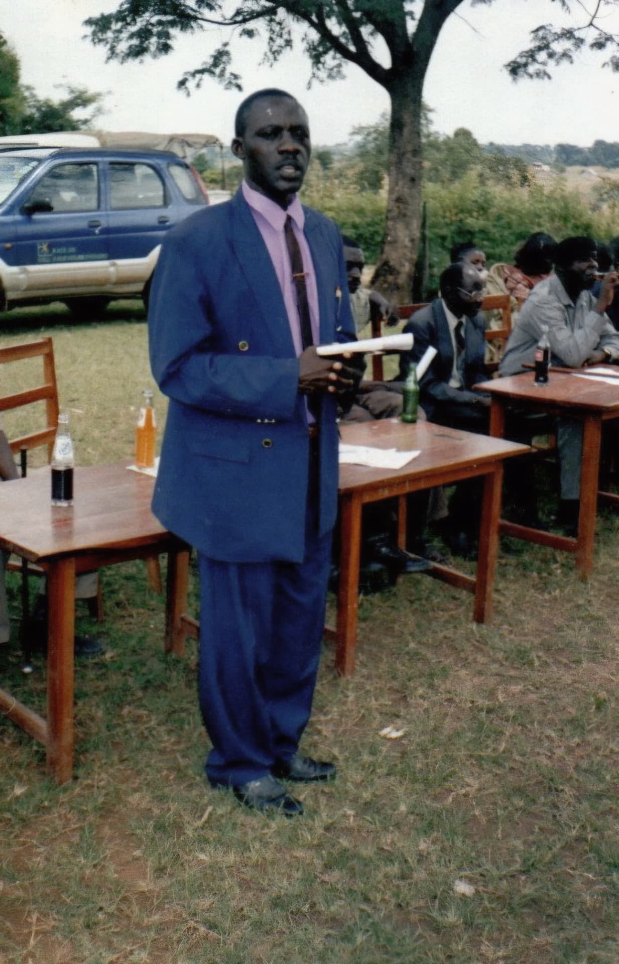 A man in a blue suit addressing officials seated at outdoor tables