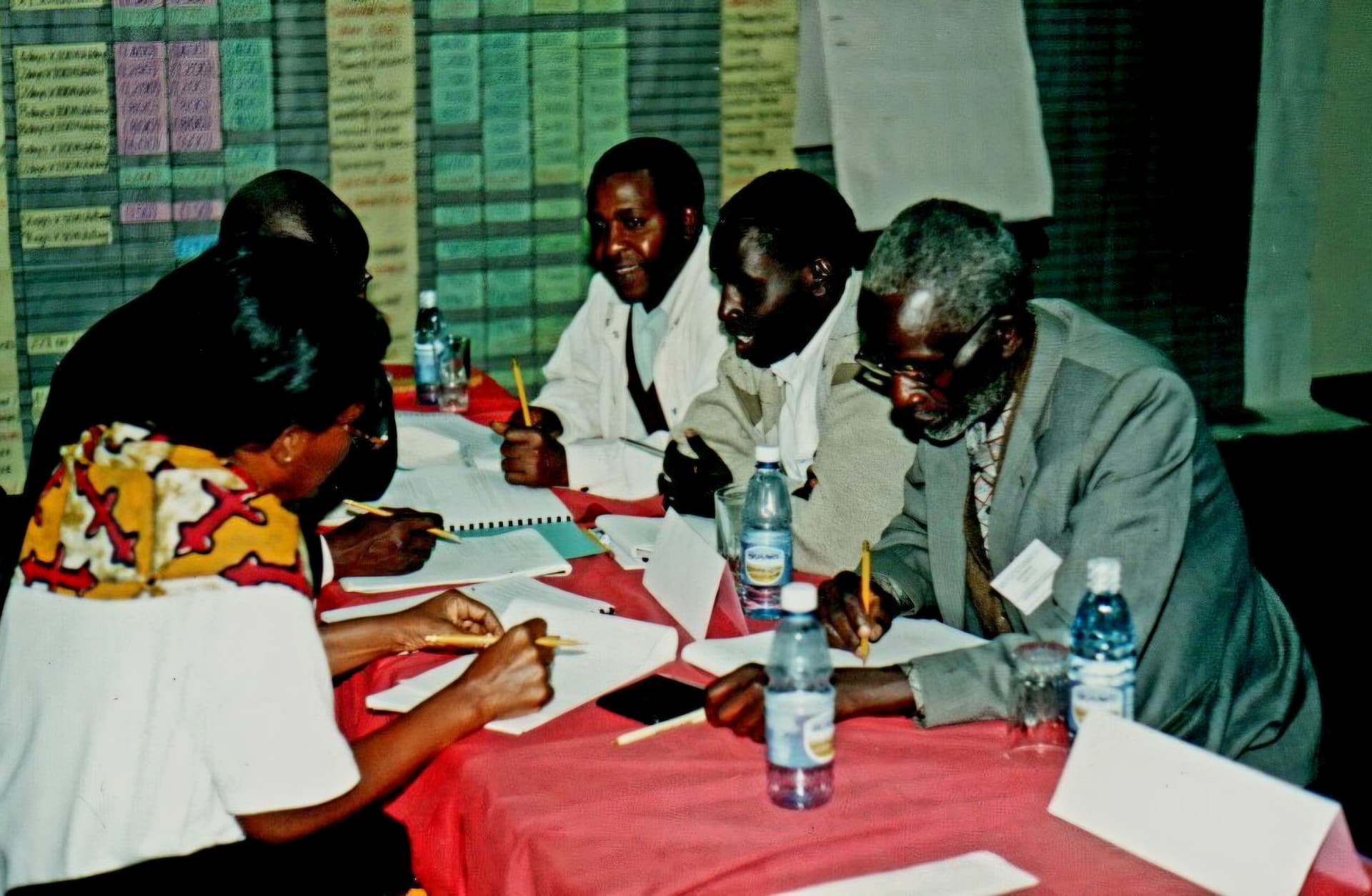 Four participants writing at a workshop table with planning charts on the wall behind them