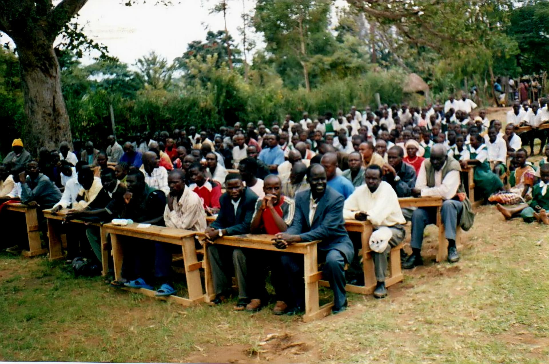 A large crowd of adults and schoolchildren seated on wooden benches under trees