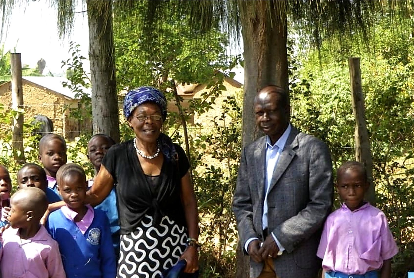 Alice Nabwera standing with schoolchildren and a school official under trees at Chemwa Primary School