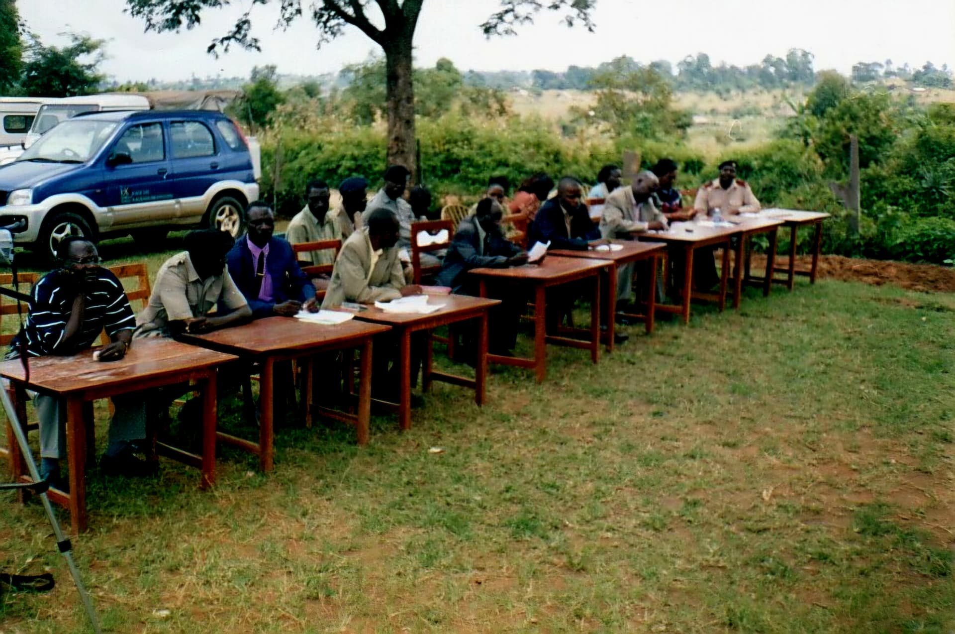 A row of officials seated at wooden tables outdoors during a project meeting