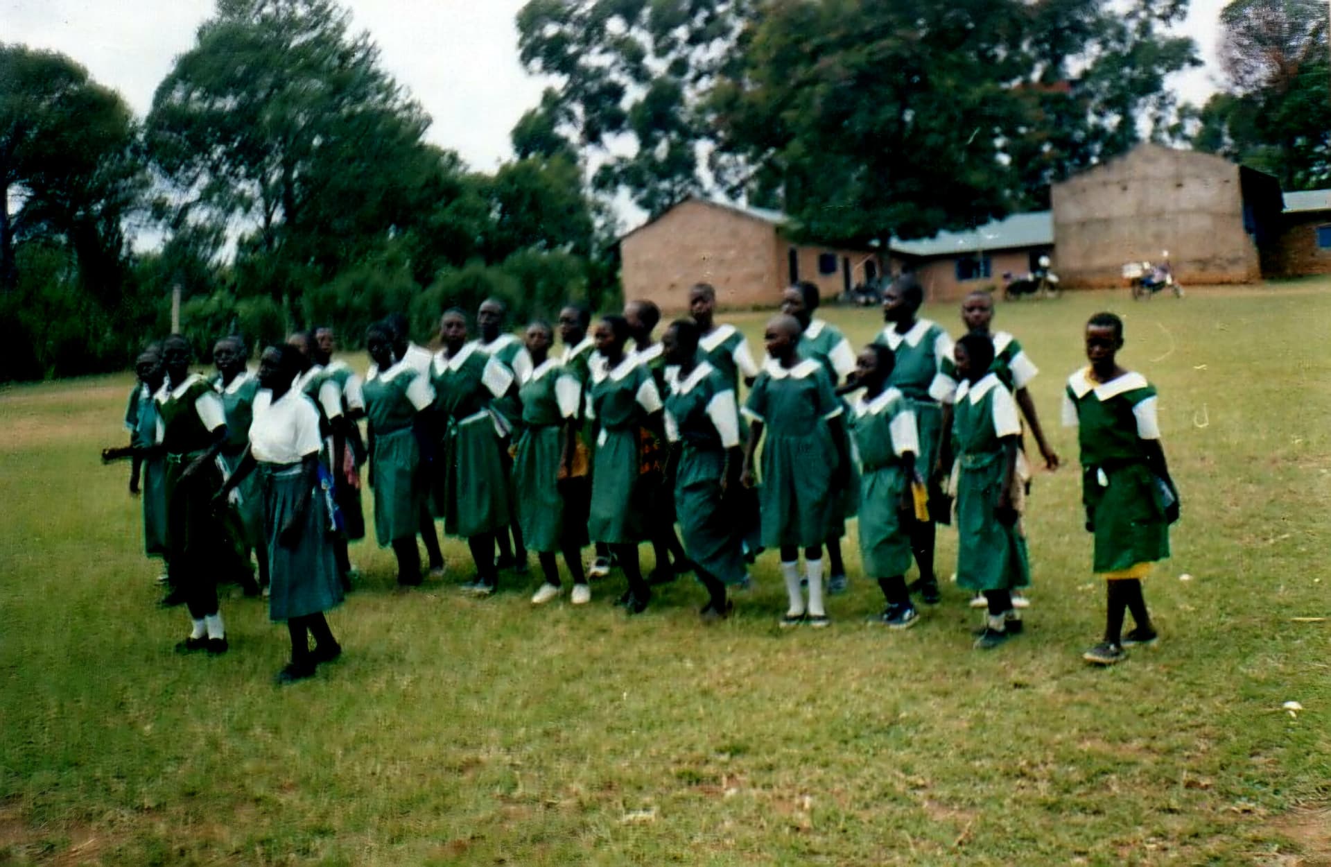 A column of pupils in green uniform standing on a school field with classrooms behind