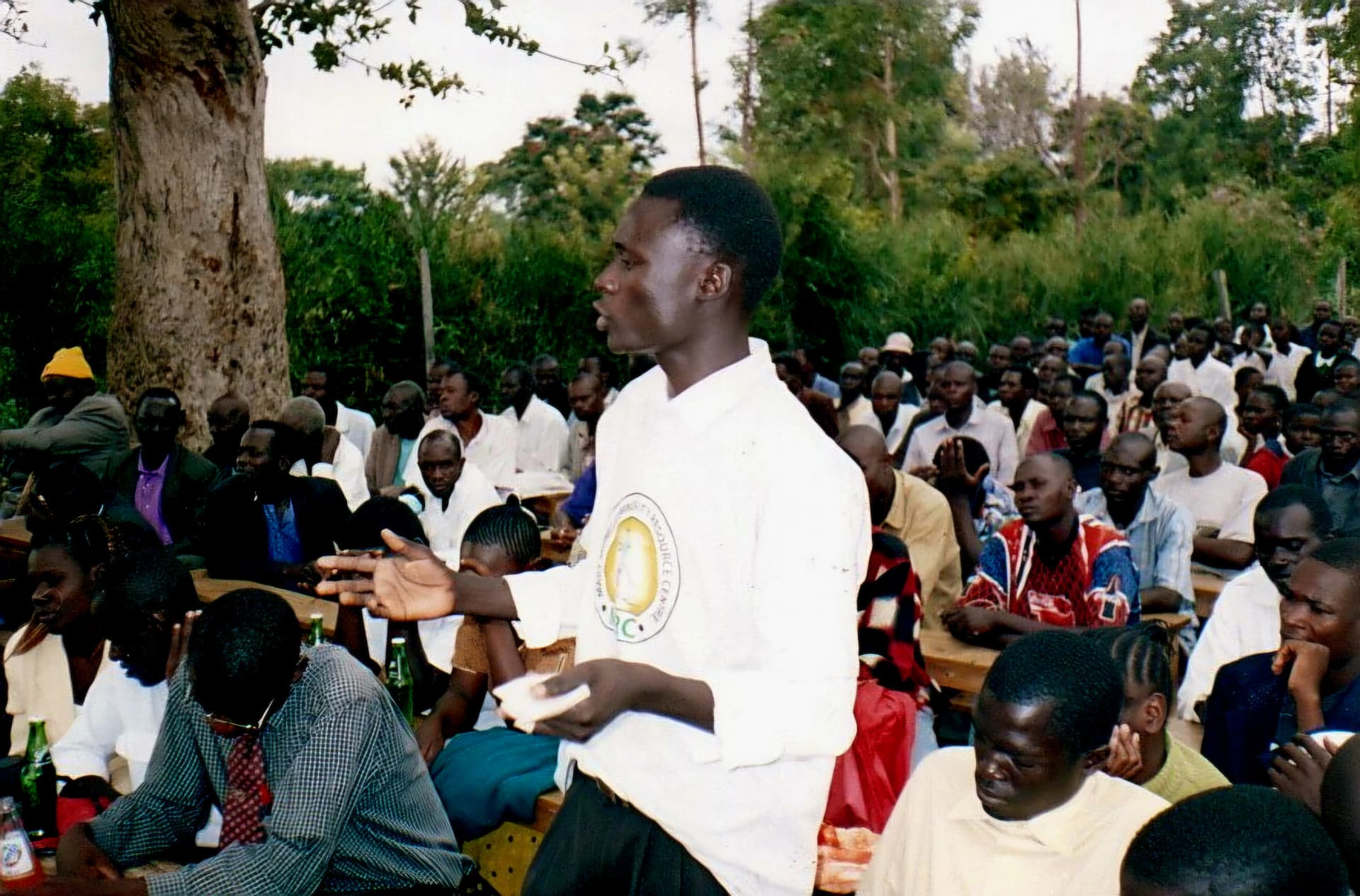 A young man in an MCRC branded shirt speaking while standing among a seated crowd