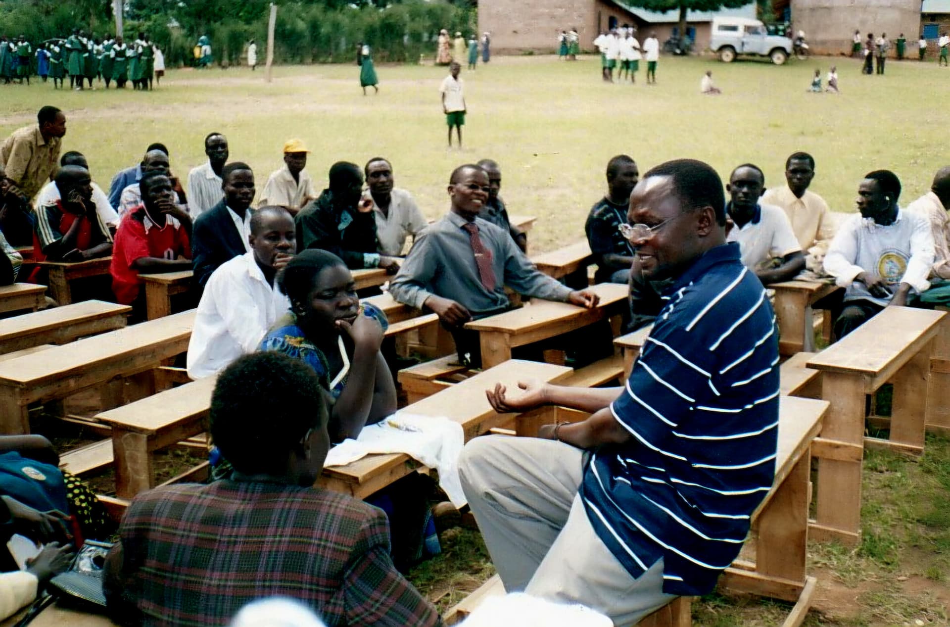 A man seated on a desk in conversation with community members on a school field