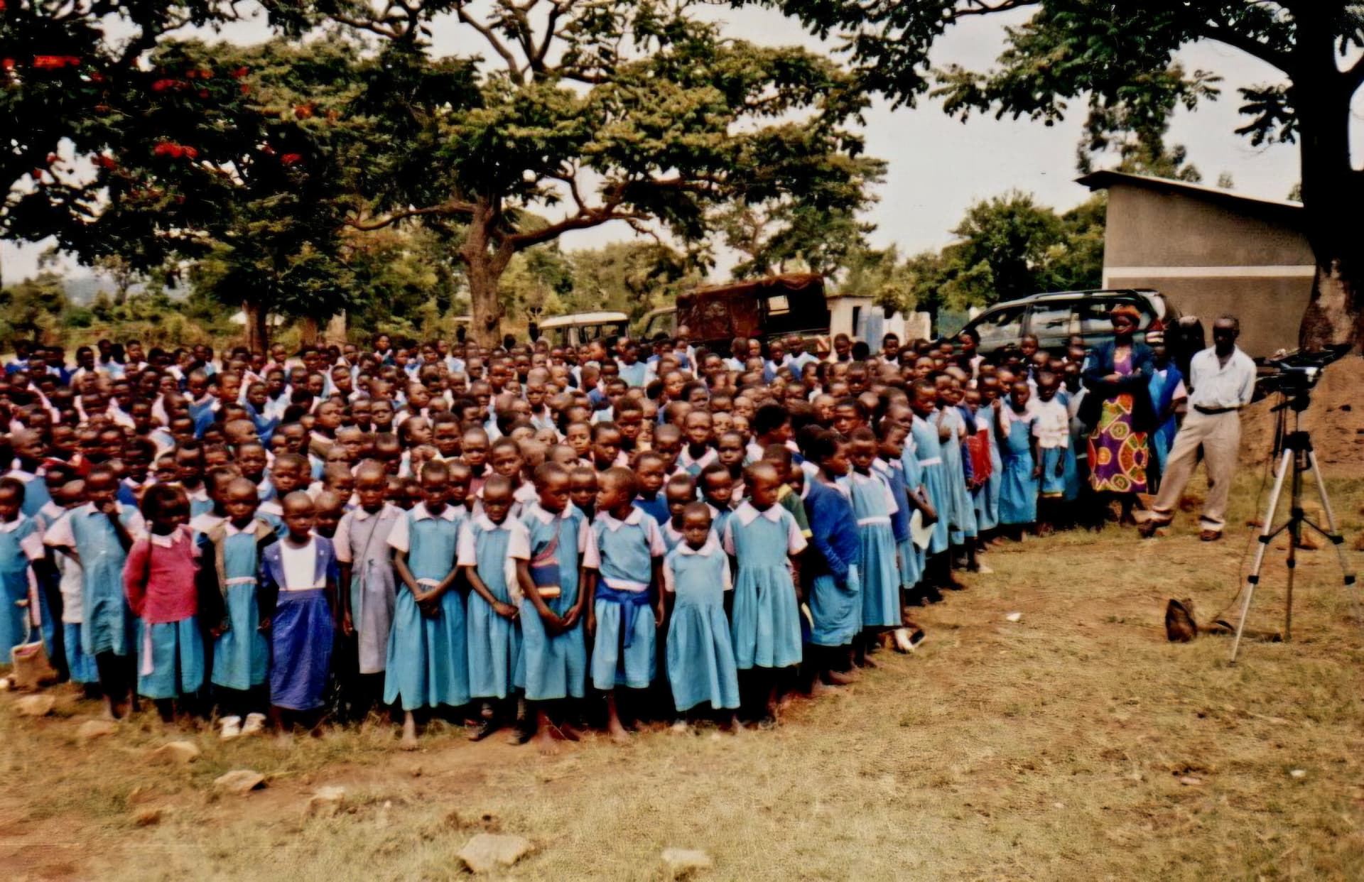 Hundreds of pupils in blue uniform assembled under trees in a school compound